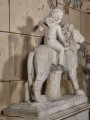 Amorek na lwie. Rzeźba marmurowa przedstawia kroczącego lwa z siedzącym na niej amorkiem.  Widok od tyłu na tle kamiennej ściany.