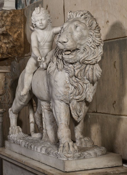 Amorek na lwie. Rzeźba marmurowa przedstawia kroczącego lwa z siedzącym na niej amorkiem. Widok od strony głowy lwa na tle kamiennej ściany.