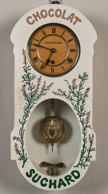 Zegar wiszący. ﻿Bardzo oryginalny zegar reklamowy firmy Suchard. Ścianka frontowa skrzynki zegarowej zdobiona jest motywem lawendy i napisem CHOCOLAT SUCHARD. W ściance znajdują się dwa otwory, z których górny pozwala obserwować białą, emaliowaną tarczę zegara z wykonanymi w kolorze czarnym cyframi rzymskimi. Poniżej znajduje się owalny otwór, przez który widoczna jest praca wahadła.