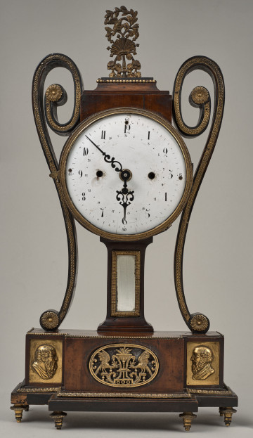 Zegar stojący z dużą białą tarczą. Wykonany z drewna ze zdobieniami z brązu. Obudowa zegara posiada zarys stylizowanego wazonu z esowato wygiętymi uchami.