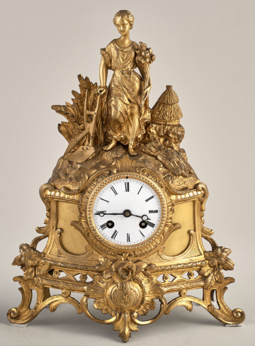 Zegar kominkowy z brązu złoconego. Zegar zdobiony motywami roślinnymi. W zwieńczeniu widoczna postać kobiety. Tarcza biała z czarnymi rzymskimi cyframi.