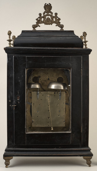 Zegar szafkowy. Ujęcie tylnej strony. Prostopadłościenna obudowa drewniana, czerniona z oszklonym otworem ukazującym mechanizm. W zwieńczeniu szafki znajduje się antabka ułatwiająca przenoszenie  zegara.