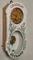 Zegar wiszący. ﻿Bardzo oryginalny zegar reklamowy firmy Suchard. Ścianka frontowa skrzynki zegarowej zdobiona jest motywem lawendy i napisem CHOCOLAT SUCHARD. W ściance znajdują się dwa otwory, z których górny pozwala obserwować białą, emaliowaną tarczę zegara z wykonanymi w kolorze czarnym cyframi rzymskimi. Poniżej znajduje się owalny otwór, przez który widoczna jest praca wahadła. Przód 3/4