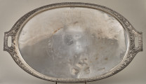 Taca srebrna, owalna z dekoracyjnym brzegiem z dwoma uchwytami ustawiona na drucianej podstawce.