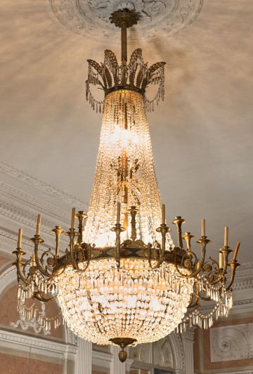 Żyrandol kryształowy z obręczą ze świeczkami w kształcie gruszki, na tle sufitu z włączonym oświetleniem.
