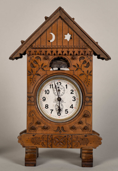 Przód zegara w formie drewnianej chatki na dwóch wspornikach z dwuspadowym dachem. W środkowej części biała tarcza zegarowa z czarnymi wskazówkami i oznaczeniem godzin.