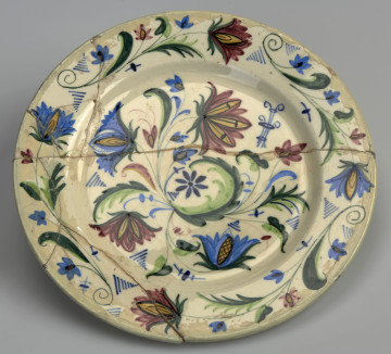 Ujęcie przodu. Płaski okrągły talerz wykonany z fajansu, pomalowany na biało i ozdobiony motywem florystycznym w kolorach: zielonym, niebieskim, różowym i żółtym.