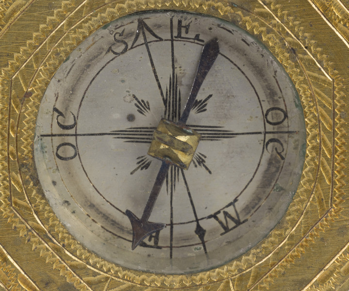 zbliżenie na ośmiokątną tarczę grawerowaną w pasy dekoracyjnej bordiury oraz przeszkolną mosiężną puszkę z kompasem