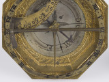zbliżenie na ośmiokątną tarczę grawerowaną w pasy dekoracyjnej bordiury oraz przeszkolną mosiężną puszkę z kompasem