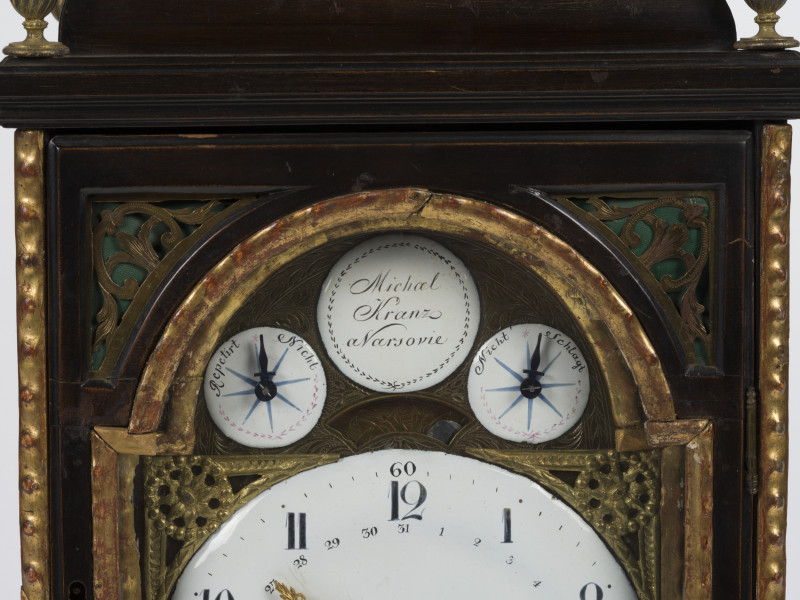 zbliżenie na tarczę zegara ze wskazaniami kalendarza, dni miesiąca, godzinami, kwadransami, minutami i sekundami, z repetierem oraz drewnianą, przeszkloną obudowę z dekoracyjnymi ażurami akustycznymi z mosiądzu grawerowanego
