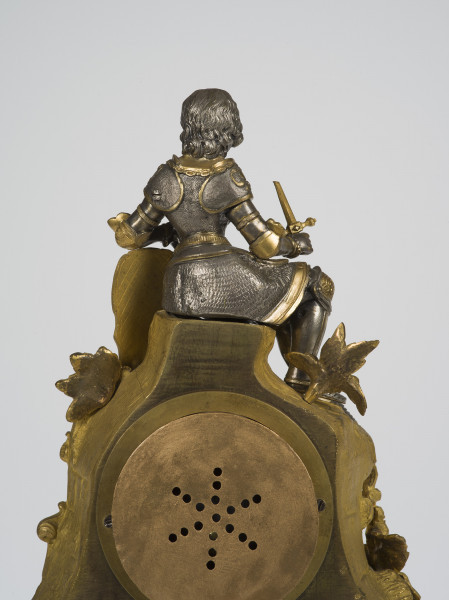 zbliżenie na zwieńczenie zegara z tyłu - figurka siedzącej na kamieniu Joanny d'Arc w zbroi