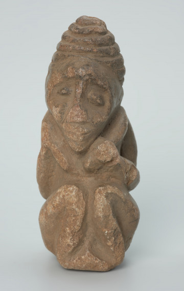 rzeźba; przedmiot obrzędowy; Figura kultu zmarłych - Ujęcie z przodu. Rzeźbiona w szarobeżowym steatycie siedząca postać ludzka z przykurczonymi nogami i dużą głową o podłużnej, trójkątnej twarzy, której dłonie spoczywają na kolanach tuż pod brodą.