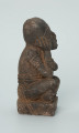 Ujęcie z boku z prawej. Rzeźbiona w grafitowym kamieniu postać ludzka - najprawdopodobniej mężczyzna - w pozycji siedzącej z nogami skrzyżowanymi. Rzeźba ma charakterystyczne bransolety – dwie na ramionach oraz jedną na nadgarstku - które ozdobiono wzorem w postaci ukośnych linii. Widoczne ślady czerwonego piasku, rysy, mikropęknięcia oraz ubytek z tyłu głowy.