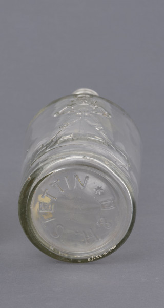 szklana butelka z szerokim korpusem - Ujęcie z dołu; Butelka piwa z grubego półbiałego przeźroczystego szkła. Korpus szeroki, lekko zwężający się ku dołowi, a od góry przechodzący w szyjkę zakończoną płaskimi pierścieniem. Czytelne szwy boczne.