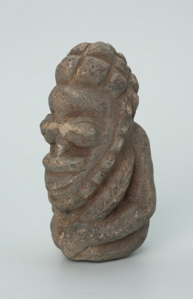 rzeźba; przedmiot obrzędowy; Figura kultu zmarłych - Ujęcie z przodu z lewej strony. Rzeźbiona w szarobeżowym steatycie siedząca postać ludzka o cechach męskich, w prawej dłoni trzymajaca przedmiot przypominający grot, a w lewej pióro lub liść.