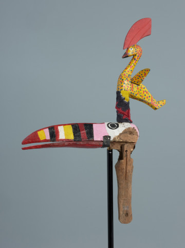 Ujęcie z boku a lewej strony. Kolorowy ptak zrobiony z drewna z ruchomą żuchwą poruszaną za pomocą sznurka. Długi, pomalowany w kolorowe paski dziób. Na głowie umieszczony drugi, mniejszy ptak w żółtym kolorze z czerwonym czubem.