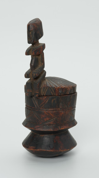 drewninany pojemnik na tytoń z pokrywką - Ujęcie z przodu, z prawej strony. Drewniany pojemnik na tytoń z pokrywą, na której znajduje się rzeźbiona, siedząca postać.