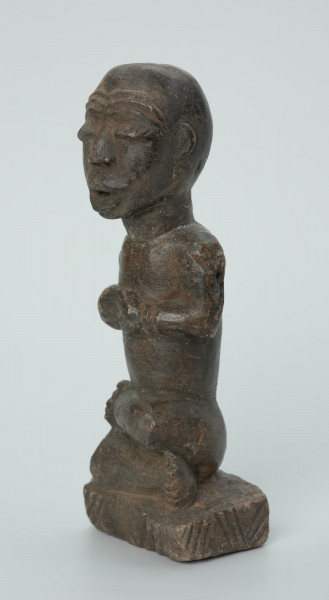 Ujęcie z przodu z lewej. Rzeźbiona w grafitowym kamieniu postać ludzka w pozycji siedzącej - najprawdopodobniej mężczyzna. Rzeźba ma charakterystyczne nacięcia - skaryfikacje wykonane na ramionach oraz plecach. Widoczne rysy i mikropęknięcia.