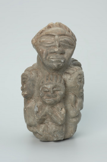 rzeźba; przedmiot obrzędowy; figura kultu zmarłych - Ujęcie z przodu. Rzeźba o gładkiej powierzchni w szarym steatycie, przedstawiająca postać ludzką o cechach kobiecych, otoczoną piątką mniejszych, dziecięcych sylwetek w różnych pozach.