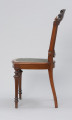 krzesło - Ujęcie z lewej strony. Krzesło z płasko wyściełanym siedziskiem i oparciem z plecionki z rafii. Przednie nogi toczone, tralkowe, w części środkowej wydzielonej wałkami - kanelowane. Tylne nogi o przekroju okrągłym, lekko wygięte, przechodzące w prostokątne słupki zaplecka. Siedzenie w kształcie podkowiastym, z przodu lekko zaokrąglone na zewnątrz, w środku wyściełane - tkanina mocowana pinezkami tapicerskimi. Na styku przednich nóg i ramy siedziska ozdobna kostka z płycinami na obu ściankach zewnętrznych. Rama zaplecka w formie medalionu, wypełnionego plecionką z rafii. Słupki zaplecka dekorowane wgłębnym reliefem w formie płycin i spirali, a łuk zwieńczenia zdobi dekoracja o formach rollwerkowo-roślinnych. Ponad kostkami wieńczącymi boczne ramy zaplecka toczone gałki. Siedzisko wyściełane tkaniną w kolorze zielonym (turkusowym?), z brzegami wykończonymi pasmanterią.