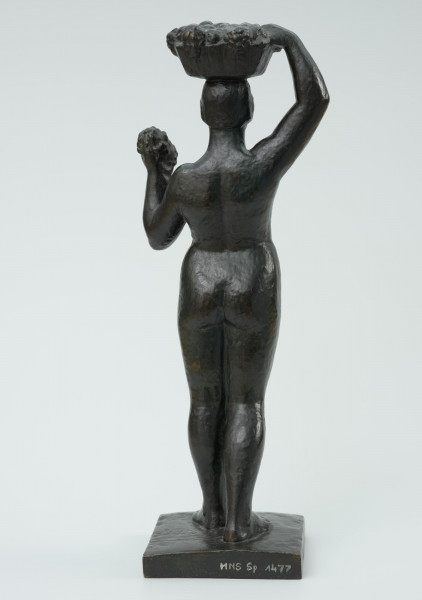 akt kobiecy - Ujęcie z tyłu; Posążek przedstawia postać stojącej kobiety o pełnych kształtach dźwigającą kosz na głowie. Sylwetka wyprostowana o masywnych nogach, biodrach i niewielkich piersiach, zupełnie naga, z okrągłą twarzą i krótkich włosach- prawa noga lekko cofnięta, lewa ręka zgięta w łokciu dzierży na wysokości barku kiść owoców, prawa uniesiona ręka przytrzymuje na głowie płaski okrągły niski kosz wypełniony owocami. Figurka stoi na kwadratowej niskiej podstawce.