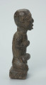 figura kultu przodków - Ujęcie z boku z prawej. Rzeźbiona w grafitowym kamieniu postać ludzka w pozycji siedzącej - najprawdopodobniej mężczyzna. Rzeźba ma charakterystyczne nacięcia - skaryfikacje wykonane na ramionach oraz plecach. Widoczne rysy i mikropęknięcia.
