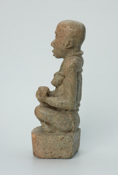 figura kultu przodków - Ujęcie z boku z lewej. Rzeźbiona w biało-szarym kamieniu dość duża postać kobiety w pozycji siedzącej z nogami skrzyżowanymi. Kobieta siedzi na małej, kwadratowej, lekko zaokrąglonej na rogach podstawie, w dłoniach trzyma dziecko. Widoczne rysy, mikropęknięcia i liczne malutkie otworki.