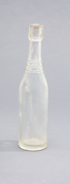 szklana butelka z wyodrębnioną szyjką z trzema pierścieniami - Ujęcie z tyłu; Szklana butelka piwówka z grubego szkła butelkowego półbiałego przezroczystego. Szyjka wyodrębniona trzema wypukłymi, wąskimi pierścieniami, umieszczonymi w dolnej części szyjki. U dołu pogrubionego wylewu pogrubienia na dwa otwory do zamknięcia pałąkowego. Dno zagłębione płaskie. Zamknięcie pałąkowe. Ślad szwu bocznego.