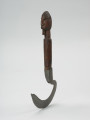 narzędzie rolnicze - Ujęcie z przodu z lewej strony. Sierp o kutym z żelaza, łukowatym ostrzu. Rączka drewniana, walcowata, zakończona rzeźbą główki z brodą w formie nacinanego liniami walca.