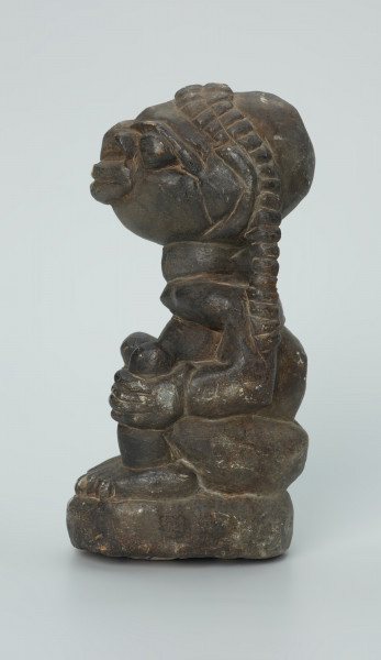 rzeźba; przedmiot obrzędowy; Figura kultu sił wegetacji - Ujęcie lewy bok. Rzeźbiony w grafitowym steatycie, na owalnej podstawie, mężczyzna w pozycji siedzącej z opaską przedstawioną za pomocą wypukłego ornamentu kostkowego, której końce opadają na ramiona i plecy postaci.