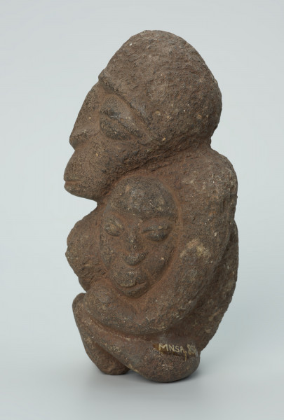 rzeźba; przedmiot obrzędowy; figura kultu zmarłych - Ujęcie lewy bok. Rzeźbiona w ziemistym steatycie siedząca postać kobiety z wyłaniającymi się symetrycznie zza jej boków dwoma ludzkimi głowami.
