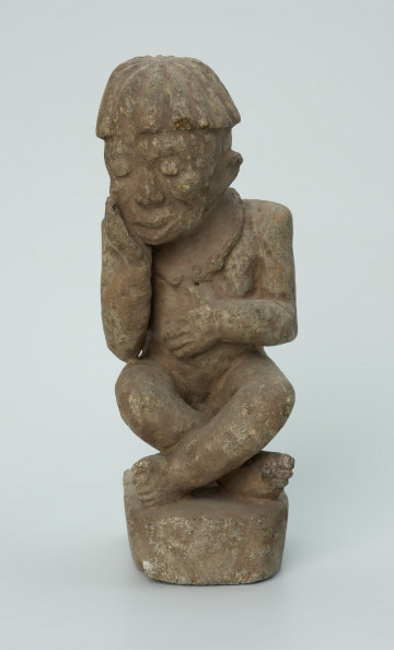 Ujęcie z przodu. Rzeźbiona w biało-szarym kamieniu postać mężczyzny w pozycji siedzącej. Mężczyzna siedzi na okrągłym, malutkim stołku, ma charakterystyczne nakrycie głowy - czapkę modelowaną promieniście oraz naszyjnik. Widoczne rysy, mikropęknięcia i malutkie otworki.