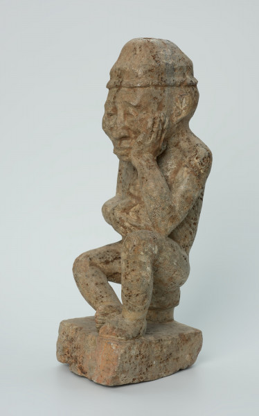 figura kultu przodków - Ujęcie przodu z lewej. Ustawiona na kwadratowej podstawie, wyrzeźbiona w biało-szarym kamieniu postać zamyślonego, siedzącego na stołku mężczyzny w czapce, o zaokrąglonych rogach i spłaszczonej części górnej.