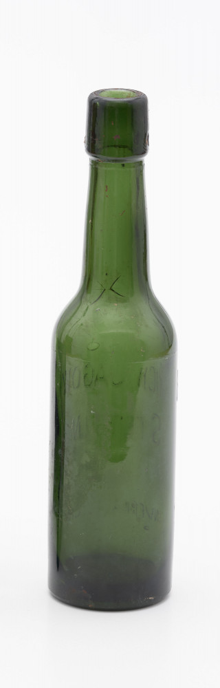szklana butelka w ciemnym kolorze oliwkowo-zielonym - Ujęcie z tyłu; Szklana butelka piwówka z grubego szkła butelkowego w ciemnym kolorze oliwkowo-zielonym. Szyjka wyodrębniona z pogrubieniem przy wylewie, u dołu pogrubienia z dwoma otworami do zamknięcia pałąkowego. Dno zagłębione płaskie. Ślad szwóww bocznych.