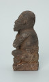 Ujęcie z boku z lewej. Rzeźbiona w grafitowym kamieniu postać ludzka - najprawdopodobniej mężczyzna - w pozycji siedzącej z nogami skrzyżowanymi. Rzeźba ma charakterystyczne bransolety – dwie na ramionach oraz jedną na nadgarstku - które ozdobiono wzorem w postaci ukośnych linii. Widoczne ślady czerwonego piasku, rysy, mikropęknięcia oraz ubytek z tyłu głowy.
