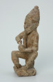 Ujęcie z przodu z lewej. Rzeźbiona w biało-szarym kamieniu postać mężczyzny palącego fajkę w pozycji siedzącej. Mężczyzna siedzi na okrągłym, malutkim stołku, ma charakterystyczne nakrycie głowy - spiczastą, ozdobioną łączącymi się ukośnymi nacięciami czapkę o zaokrąglonych rogach i pomponiku. Widoczne rysy, mikropęknięcia, liczne malutkie otworki i przebarwienia.