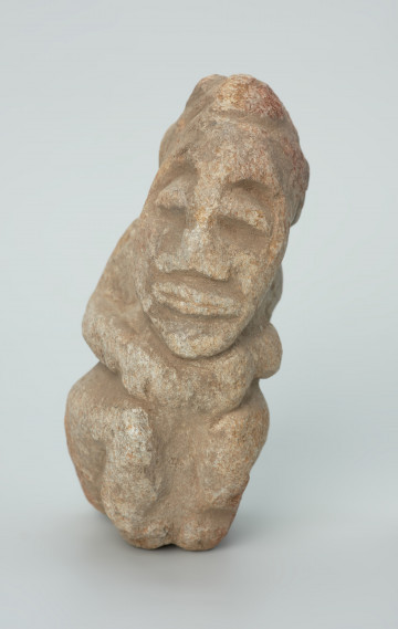 rzeźba; Figura kultu zmarłych - Ujęcie z przodu. Rzeźbiona w szarobeżowym steatycie siedząca postać ludzka z przykurczonymi nogami i dużą głową o podłużnej, trójkątnej twarzy, której dłonie spoczywają na kolanach tuż pod brodą.