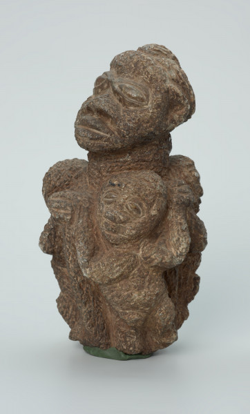 rzeźba; przedmiot obrzędowy; figura kultu zmarłych - Ujęcie z przodu z lewej strony. Rzeźbiona w ziemistym steatycie siedząca postać ludzka o cechach kobiecych, otoczona przez trójkę mniejszych, dzieciecych postaci, skierowanych ku niej z uniesionymi rękoma.