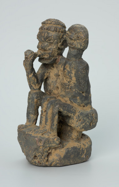 figura kultu przodków - Ujęcie z przodu z lewej. Rzeźbione w grafitowym kamieniu dwie złączone ze sobą postaci ludzkie w pozycji siedzącej. Stykają się ramionami, tułowiem oraz nogami. Jedna z nich – najprawdopodobniej mężczyzna - trzyma w prawej dłoni fajkę. Rzeźba pokryta jest piaskiem. Widoczne rysy, mikropęknięcia, malutkie otworki, przebarwienia oraz ślady klejenia w okolicach szyi.
