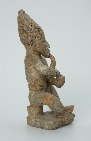 Ujęcie z boku z prawej. Rzeźbiona w biało-szarym kamieniu postać mężczyzny palącego fajkę w pozycji siedzącej. Mężczyzna siedzi na okrągłym, malutkim stołku, ma charakterystyczne nakrycie głowy - spiczastą, ozdobioną łączącymi się ukośnymi nacięciami czapkę o zaokrąglonych rogach i pomponiku. Widoczne rysy, mikropęknięcia, liczne malutkie otworki i przebarwienia.