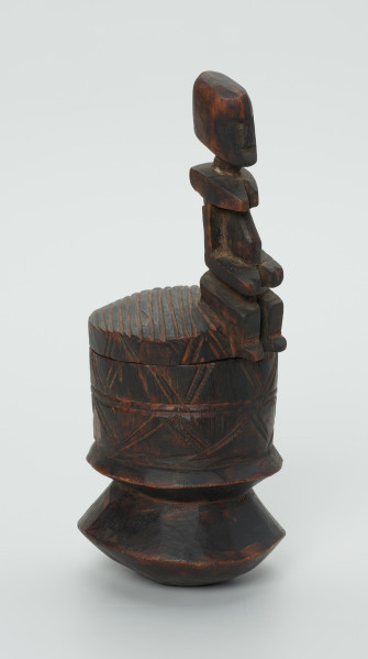 drewninany pojemnik na tytoń z pokrywką - Ujęcie z boku. Drewniany pojemnik na tytoń z pokrywą, na której znajduje się rzeźbiona, siedząca postać.