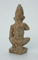 Ujęcie z tyłu. Rzeźbiona w biało-szarym kamieniu postać mężczyzny palącego fajkę w pozycji siedzącej. Mężczyzna siedzi na okrągłym, malutkim stołku, ma charakterystyczne nakrycie głowy - spiczastą, ozdobioną łączącymi się ukośnymi nacięciami czapkę o zaokrąglonych rogach i pomponiku. Widoczne rysy, mikropęknięcia, liczne malutkie otworki i przebarwienia.