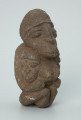 rzeźba; przedmiot obrzędowy; figura kultu zmarłych - Ujęcie z przodu z prawej strony. Rzeźbiona w ziemistym steatycie siedząca postać kobiety z wyłaniającymi się symetrycznie zza jej boków dwoma ludzkimi głowami.