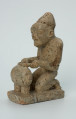 Ujęcie z przodu z lewej. Rzeźbiona w szarym kamieniu postać mężczyzny w pozycji klęczącej, grającego na bębnie szczelinowym tam-tamie. Widoczne rysy, mikropęknięcia, liczne malutkie otworki, przebarwienia, ślady klejenia oraz ubytek na lewym łokciu.