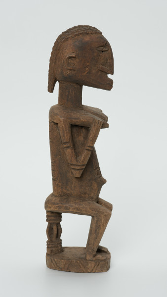 drewniana, rzeźbiona figura - Ujęcie z lewego boku. Drewniana, rzeźbiona figura kobiety siedzącej na stołku.