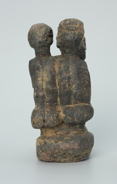 figura kultu przodków - Ujęcie z tyłu. Rzeźbione w grafitowym kamieniu dwie złączone ze sobą postaci ludzkie w pozycji siedzącej. Stykają się ramionami, tułowiem oraz nogami. Jedna z nich – najprawdopodobniej mężczyzna - trzyma w prawej dłoni fajkę. Rzeźba pokryta jest piaskiem. Widoczne rysy, mikropęknięcia, malutkie otworki, przebarwienia oraz ślady klejenia w okolicach szyi.