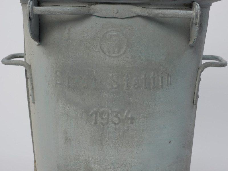 metalowy pojemnik z pokrywą - detal; Na korpusie z przodu napis Stadt Stettin oraz data pod napisem 1934. Widoczne boczne uchwyty.