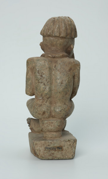 Ujęcie z tyłu. Rzeźbiona w biało-szarym kamieniu postać mężczyzny w pozycji siedzącej. Mężczyzna siedzi na okrągłym, malutkim stołku, ma charakterystyczne nakrycie głowy - czapkę modelowaną promieniście oraz naszyjnik. Widoczne rysy, mikropęknięcia i malutkie otworki.