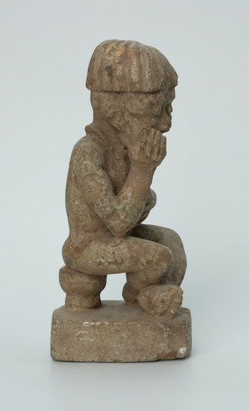 Ujęcie z boku z prawej. Rzeźbiona w biało-szarym kamieniu postać mężczyzny w pozycji siedzącej. Mężczyzna siedzi na okrągłym, malutkim stołku, ma charakterystyczne nakrycie głowy - czapkę modelowaną promieniście oraz naszyjnik. Widoczne rysy, mikropęknięcia i malutkie otworki.