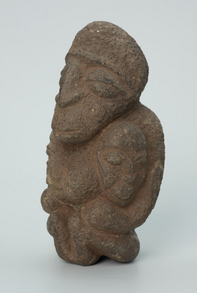 rzeźba; przedmiot obrzędowy; figura kultu zmarłych - Ujęcie z przodu z lewej strony. Rzeźbiona w ziemistym steatycie siedząca postać kobiety z wyłaniającymi się symetrycznie zza jej boków dwoma ludzkimi głowami.